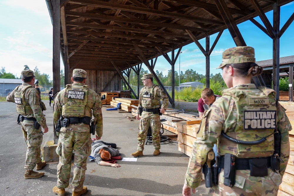 The image shows U.S. Military Police personnel conducting what appears to be a training exercise at an outdoor covered facility. Several soldiers in OCP (Operational Camouflage Pattern) uniforms with 'Military Police' tactical vests are present, with one individual lying on the ground as part of the scenario. Name tapes visible include 'CAMACHO' and 'MOODY', suggesting this is a documented training event.