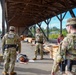 The image shows U.S. Military Police personnel conducting what appears to be a training exercise at an outdoor covered facility. Several soldiers in OCP (Operational Camouflage Pattern) uniforms with 'Military Police' tactical vests are present, with one individual lying on the ground as part of the scenario. Name tapes visible include 'CAMACHO' and 'MOODY', suggesting this is a documented training event.