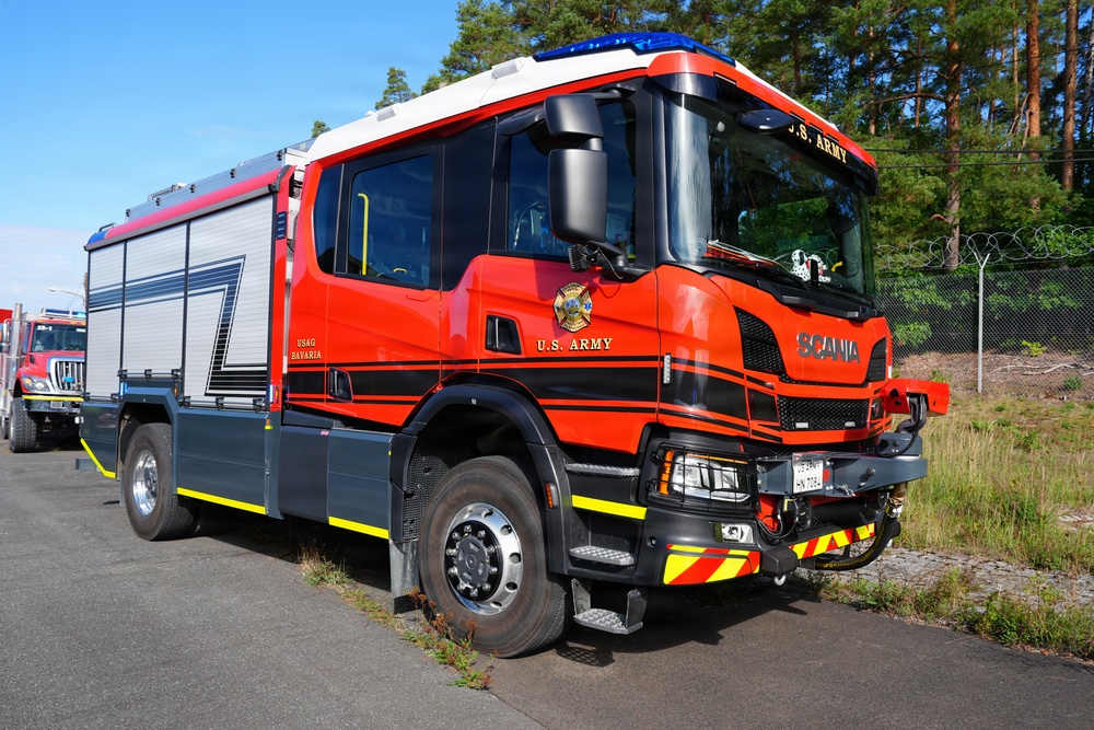 A bright red Scania fire truck belonging to the U.S. Army, specifically USAG Bavaria, is parked on a paved surface near a tree line and chain-link fence. The vehicle is a modern emergency response apparatus featuring blue emergency lights on the roof, yellow chevron markings on the bumper, and aluminum equipment storage compartments. The truck appears to be in excellent, near-new condition.