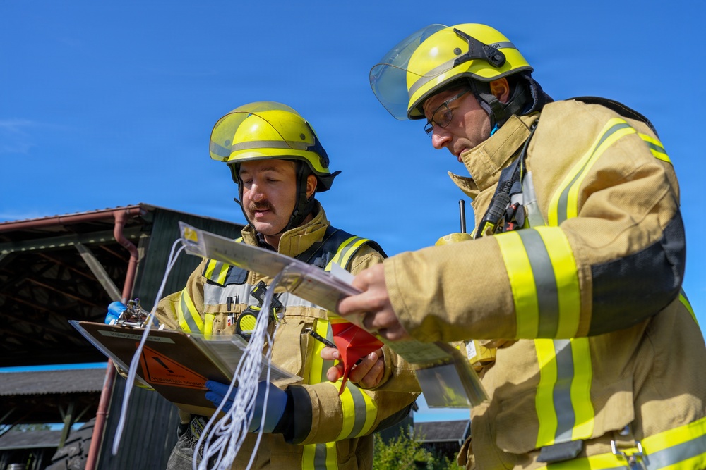 Two firefighters in full protective gear are reviewing documents and materials outdoors on a clear sunny day. They appear to be consulting reference materials, possibly during an incident response or training exercise. One firefighter holds a clipboard with a hazardous materials placard visible, while the other holds additional documents.