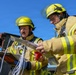 Two firefighters in full protective gear are reviewing documents and materials outdoors on a clear sunny day. They appear to be consulting reference materials, possibly during an incident response or training exercise. One firefighter holds a clipboard with a hazardous materials placard visible, while the other holds additional documents.