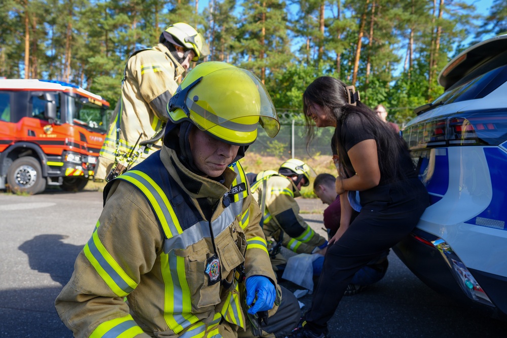 The image shows firefighters in full protective gear responding to what appears to be a vehicle incident near a car. Multiple responders in yellow high-visibility turnout gear are attending to a civilian woman with dark hair near the rear of a white vehicle. A red fire truck is visible in the background among tall pine trees.