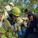 The image shows firefighters in full protective gear responding to what appears to be a vehicle incident near a car. Multiple responders in yellow high-visibility turnout gear are attending to a civilian woman with dark hair near the rear of a white vehicle. A red fire truck is visible in the background among tall pine trees.