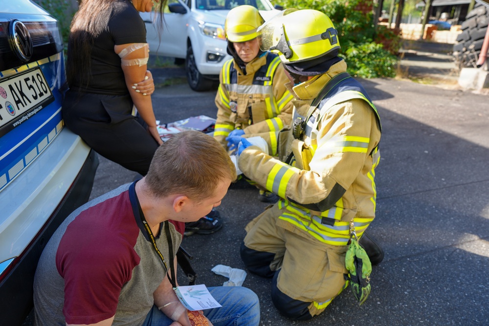 Two firefighters in yellow high-visibility gear and helmets are providing first aid to an injured person near a vehicle. A civilian man wearing a lanyard is crouching nearby, appearing to observe or assist. The scene appears to be an emergency response situation, possibly a traffic accident or training exercise, taking place on a paved outdoor area.