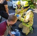 Two firefighters in yellow high-visibility gear and helmets are providing first aid to an injured person near a vehicle. A civilian man wearing a lanyard is crouching nearby, appearing to observe or assist. The scene appears to be an emergency response situation, possibly a traffic accident or training exercise, taking place on a paved outdoor area.