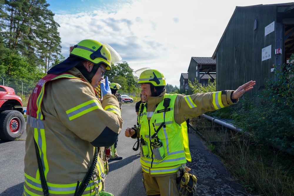 Two firefighters in high-visibility yellow-green gear and helmets appear to be communicating and coordinating during what looks like an emergency response or training exercise. One firefighter is gesturing with an outstretched arm, possibly directing personnel or indicating a direction. The scene takes place on a paved road near dark wooden structures surrounded by trees.
