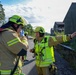 Two firefighters in high-visibility yellow-green gear and helmets appear to be communicating and coordinating during what looks like an emergency response or training exercise. One firefighter is gesturing with an outstretched arm, possibly directing personnel or indicating a direction. The scene takes place on a paved road near dark wooden structures surrounded by trees.