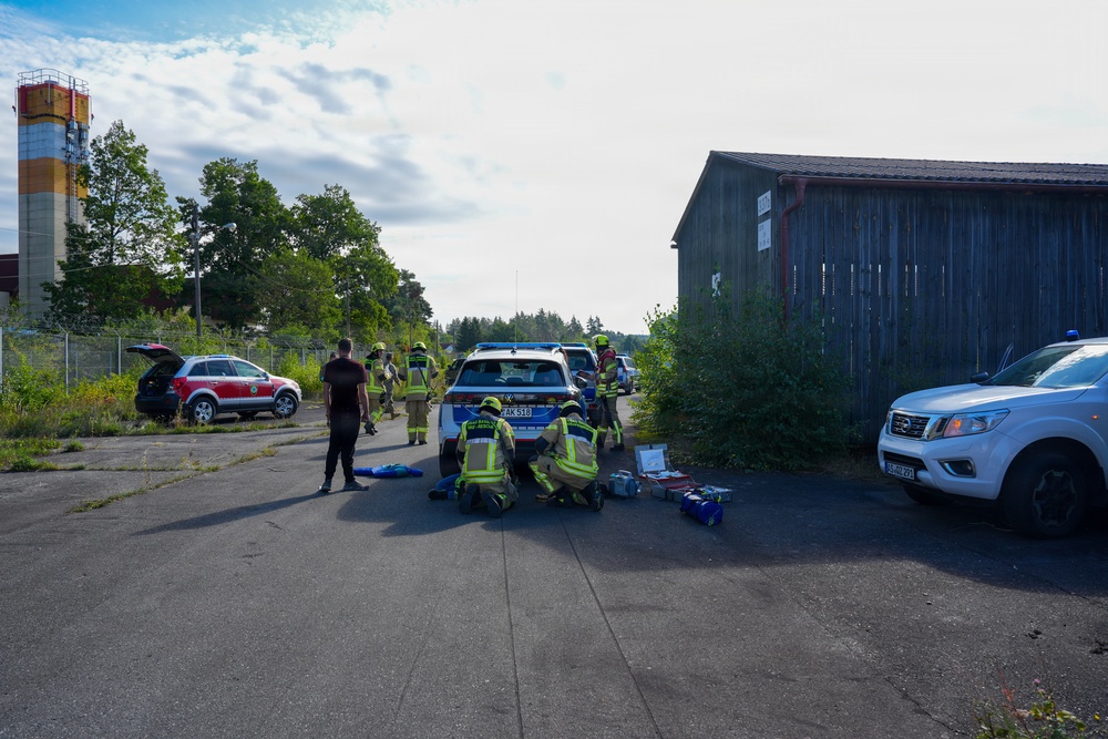 The image shows what appears to be an emergency response exercise or incident scene in an industrial or semi-abandoned area. Several personnel in high-visibility firefighter/rescue gear are gathered around vehicles, with some kneeling on the ground, possibly attending to equipment or a simulated casualty. The scene includes multiple response vehicles, a wooden barn-like structure, and an industrial tower in the background.