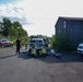The image shows what appears to be an emergency response exercise or incident scene in an industrial or semi-abandoned area. Several personnel in high-visibility firefighter/rescue gear are gathered around vehicles, with some kneeling on the ground, possibly attending to equipment or a simulated casualty. The scene includes multiple response vehicles, a wooden barn-like structure, and an industrial tower in the background.