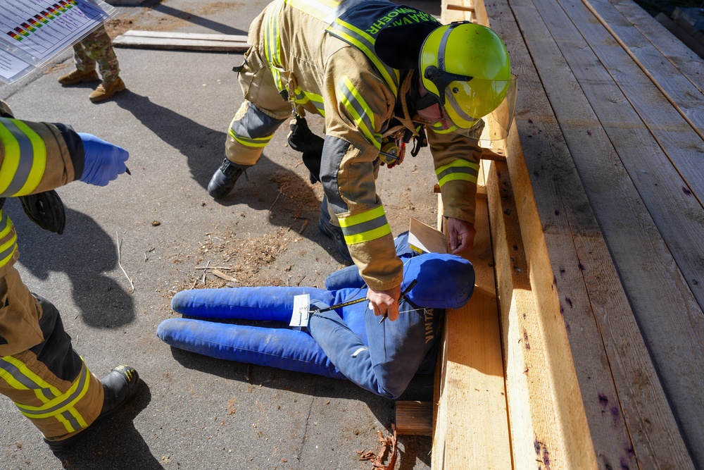 A firefighter wearing a yellow helmet and high-visibility turnout gear is attending to a training mannequin dressed in blue clothing that is partially trapped under wooden beams. The scene appears to be a rescue training exercise, with multiple firefighters present and a triage tag visible on the mannequin. Sawdust and wood debris on the ground suggest a simulated structural collapse scenario.
