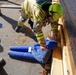 A firefighter wearing a yellow helmet and high-visibility turnout gear is attending to a training mannequin dressed in blue clothing that is partially trapped under wooden beams. The scene appears to be a rescue training exercise, with multiple firefighters present and a triage tag visible on the mannequin. Sawdust and wood debris on the ground suggest a simulated structural collapse scenario.