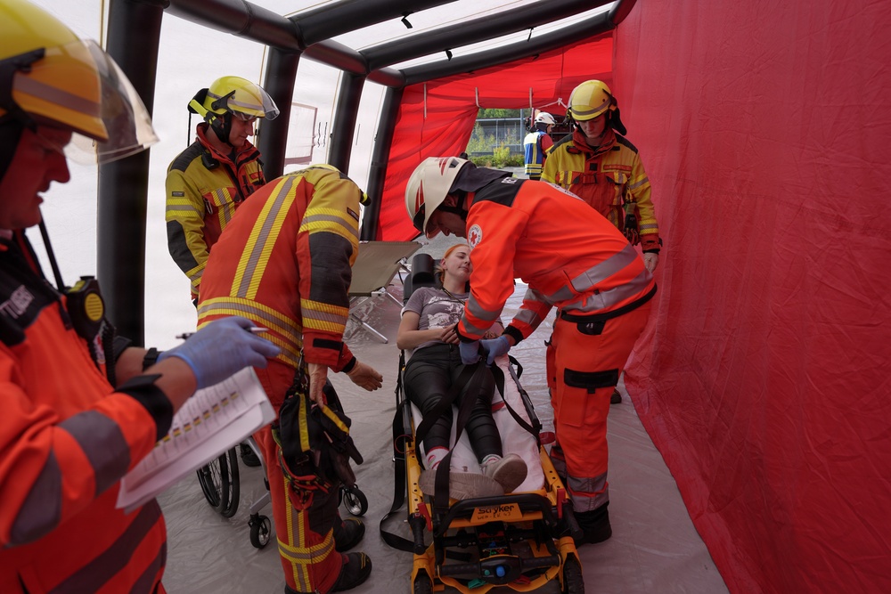 The image shows what appears to be an emergency response drill or training exercise taking place inside a red inflatable medical tent. A female patient is lying on a Stryker stretcher while multiple responders in orange and yellow high-visibility uniforms attend to her. Personnel wearing Red Cross insignia and fire brigade gear are coordinating the simulated medical response.
