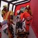 The image shows what appears to be an emergency response drill or training exercise taking place inside a red inflatable medical tent. A female patient is lying on a Stryker stretcher while multiple responders in orange and yellow high-visibility uniforms attend to her. Personnel wearing Red Cross insignia and fire brigade gear are coordinating the simulated medical response.