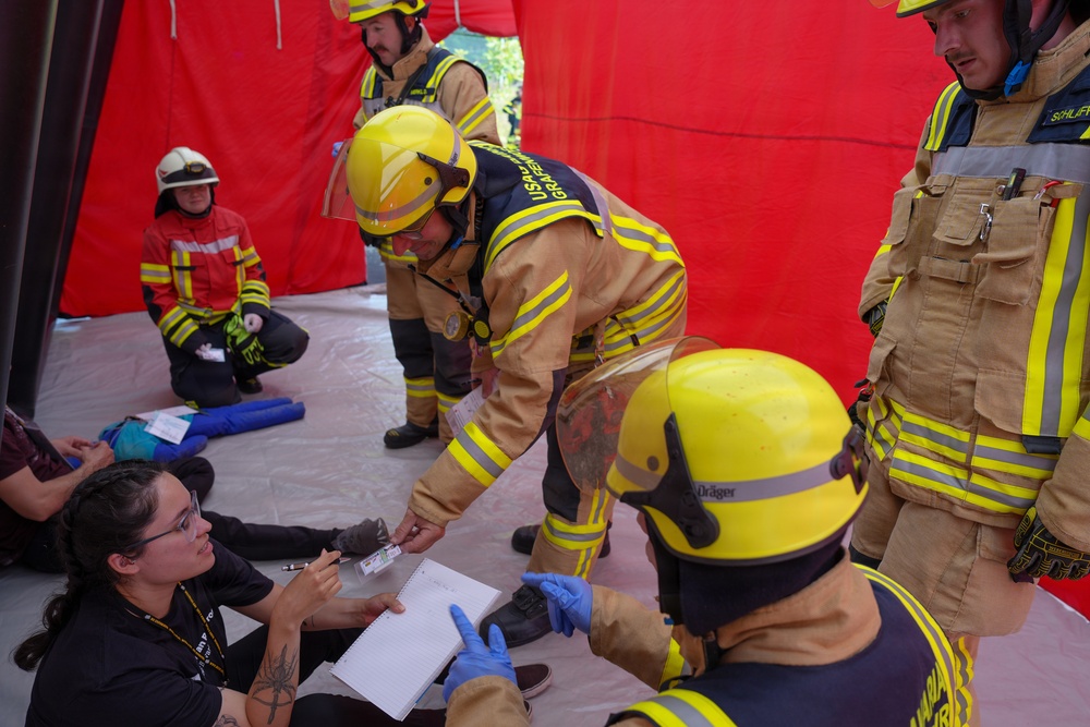 The image depicts what appears to be an emergency response training exercise or drill, taking place inside a red inflatable tent structure. Firefighters in full protective gear are interacting with a civilian woman who is seated on the ground holding a notepad, likely acting as a simulated casualty or observer. The scene suggests a mass casualty or triage exercise with multiple personnel involved.
