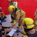 The image depicts what appears to be an emergency response training exercise or drill, taking place inside a red inflatable tent structure. Firefighters in full protective gear are interacting with a civilian woman who is seated on the ground holding a notepad, likely acting as a simulated casualty or observer. The scene suggests a mass casualty or triage exercise with multiple personnel involved.