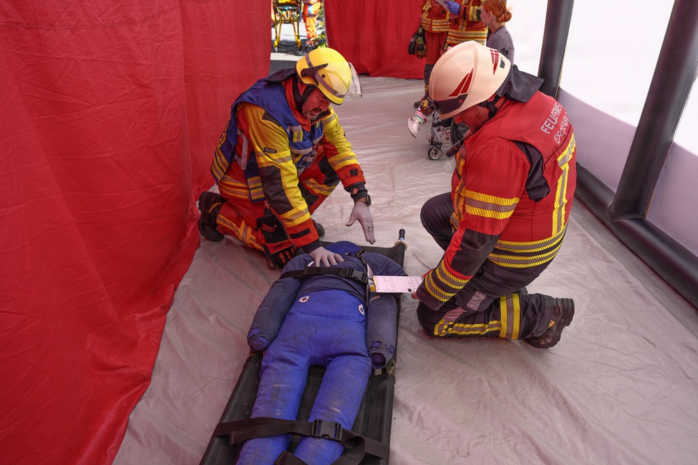 Two emergency responders in protective gear and helmets are attending to a person lying on a stretcher inside a red-walled inflatable medical tent. One responder wears a yellow and blue jacket with a yellow helmet, while the other wears a red jacket marked 'FEUERWEHR' (German for fire department) with a white helmet. This appears to be a mass casualty drill or emergency response exercise.