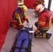 Two emergency responders in protective gear and helmets are attending to a person lying on a stretcher inside a red-walled inflatable medical tent. One responder wears a yellow and blue jacket with a yellow helmet, while the other wears a red jacket marked 'FEUERWEHR' (German for fire department) with a white helmet. This appears to be a mass casualty drill or emergency response exercise.