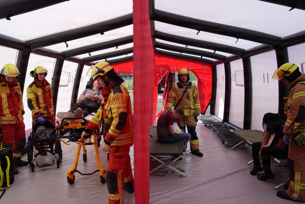 The image shows an emergency response drill or exercise taking place inside an inflatable field hospital or triage tent. Multiple firefighters and emergency medical personnel in yellow helmets and high-visibility gear are attending to simulated casualties. The scene includes a patient on a Stryker stretcher, folding cots, and several individuals acting as victims in various states of distress.