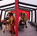 The image shows an emergency response drill or exercise taking place inside an inflatable field hospital or triage tent. Multiple firefighters and emergency medical personnel in yellow helmets and high-visibility gear are attending to simulated casualties. The scene includes a patient on a Stryker stretcher, folding cots, and several individuals acting as victims in various states of distress.
