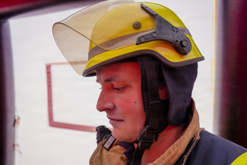 A close-up profile shot of a firefighter wearing a yellow safety helmet with a raised visor and dark ear/neck protection. The individual's face appears flushed and slightly sweaty, suggesting recent physical exertion or heat exposure. They are dressed in standard firefighting turnout gear.