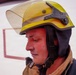 A close-up profile shot of a firefighter wearing a yellow safety helmet with a raised visor and dark ear/neck protection. The individual's face appears flushed and slightly sweaty, suggesting recent physical exertion or heat exposure. They are dressed in standard firefighting turnout gear.