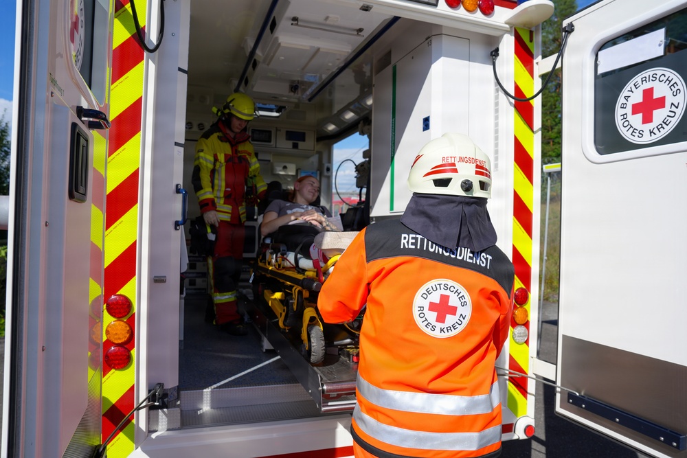 A German Red Cross (Deutsches Rotes Kreuz) emergency medical scene showing paramedics loading a patient into an ambulance. The ambulance belongs to the Bayerisches Rotes Kreuz (Bavarian Red Cross), identifiable by the logo on the vehicle door. At least two emergency responders are visible, one in an orange Rettungsdienst (rescue service) uniform and one in yellow firefighter gear, attending to a patient on a stretcher.