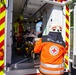 A German Red Cross (Deutsches Rotes Kreuz) emergency medical scene showing paramedics loading a patient into an ambulance. The ambulance belongs to the Bayerisches Rotes Kreuz (Bavarian Red Cross), identifiable by the logo on the vehicle door. At least two emergency responders are visible, one in an orange Rettungsdienst (rescue service) uniform and one in yellow firefighter gear, attending to a patient on a stretcher.
