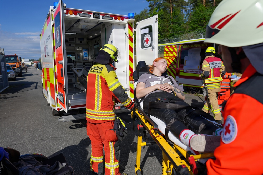 An emergency response scene showing first responders from the Grafenwöhr fire department (Feuerwehr) and Red Cross loading a patient onto a stretcher near an ambulance. The patient, a young woman with red hair, is lying on a yellow stretcher and appears conscious. Multiple emergency personnel in high-visibility gear are present, suggesting a training exercise or real incident response.
