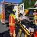 An emergency response scene showing first responders from the Grafenwöhr fire department (Feuerwehr) and Red Cross loading a patient onto a stretcher near an ambulance. The patient, a young woman with red hair, is lying on a yellow stretcher and appears conscious. Multiple emergency personnel in high-visibility gear are present, suggesting a training exercise or real incident response.