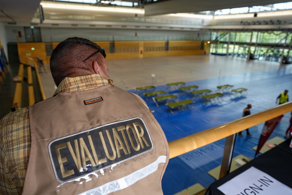 A person wearing a tan vest labeled 'EVALUATOR' on the back is observing a large indoor gymnasium or sports hall from an elevated mezzanine level. Below, the floor is set up with rows of green cots or folding tables arranged on a blue surface, suggesting an emergency shelter, disaster response drill, or mass casualty exercise. Several other individuals in safety vests are visible on the floor level.