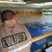 A person wearing a tan vest labeled 'EVALUATOR' on the back is observing a large indoor gymnasium or sports hall from an elevated mezzanine level. Below, the floor is set up with rows of green cots or folding tables arranged on a blue surface, suggesting an emergency shelter, disaster response drill, or mass casualty exercise. Several other individuals in safety vests are visible on the floor level.
