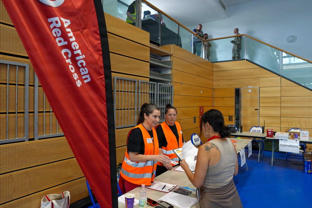 An American Red Cross relief or registration station is set up inside a modern building, likely a community center or gymnasium. Two Red Cross volunteers in orange high-visibility vests are assisting a civilian at a registration table. Additional personnel, including individuals in military uniforms, are visible on an upper mezzanine level.