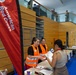 An American Red Cross relief or registration station is set up inside a modern building, likely a community center or gymnasium. Two Red Cross volunteers in orange high-visibility vests are assisting a civilian at a registration table. Additional personnel, including individuals in military uniforms, are visible on an upper mezzanine level.