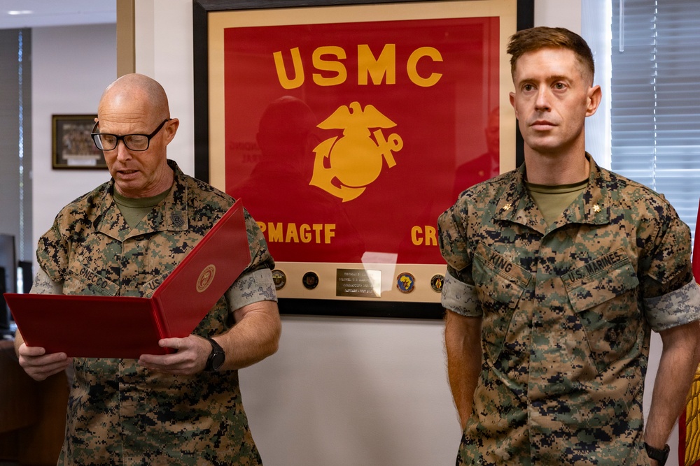 Two U.S. Marine Corps personnel in MARPAT camouflage uniforms are participating in what appears to be a formal military ceremony, likely a promotion or award presentation. The Marine on the left, identified by nametape as 'GNECCO,' is reading from a red official folder, while the Marine on the right, identified as 'KING,' stands at attention. A framed USMC Eagle, Globe, and Anchor emblem with 'RMAGTF CR' text is prominently displayed in the background.
