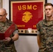 Two U.S. Marine Corps personnel in MARPAT camouflage uniforms are participating in what appears to be a formal military ceremony, likely a promotion or award presentation. The Marine on the left, identified by nametape as 'GNECCO,' is reading from a red official folder, while the Marine on the right, identified as 'KING,' stands at attention. A framed USMC Eagle, Globe, and Anchor emblem with 'RMAGTF CR' text is prominently displayed in the background.