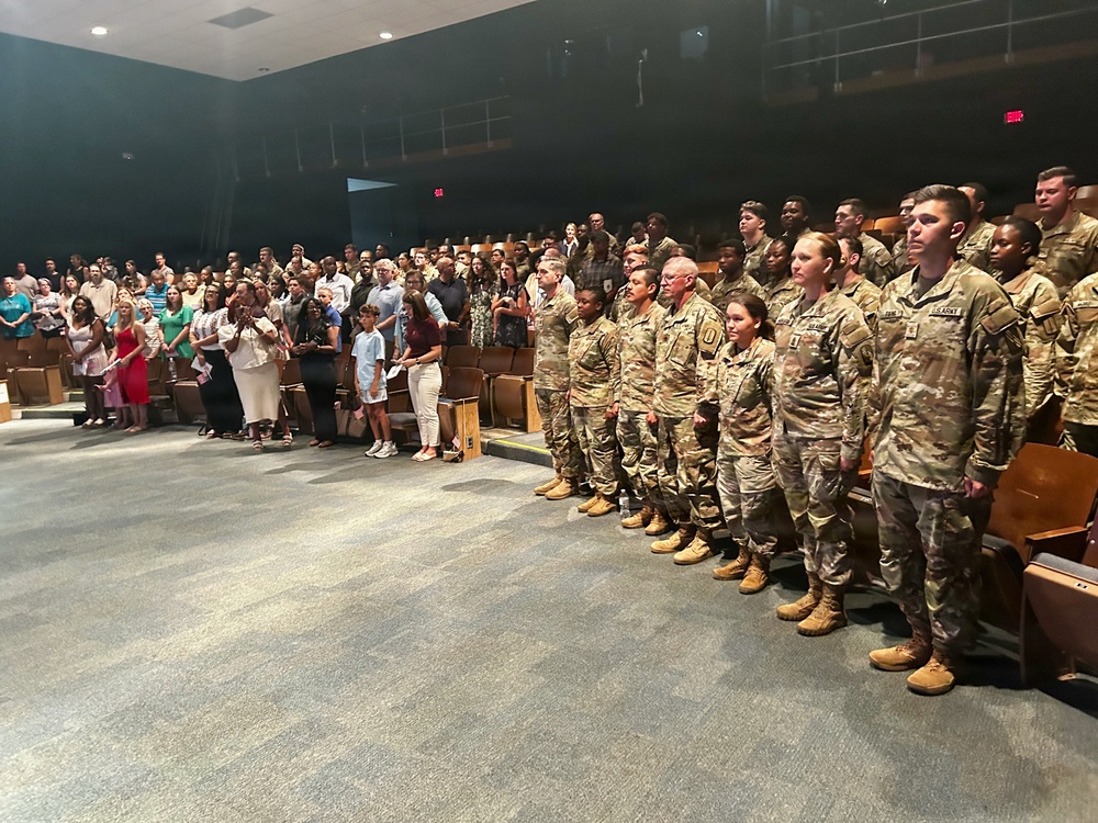 A military ceremony is taking place inside an auditorium or theater, with a formation of U.S. Army soldiers in OCP (Operational Camouflage Pattern) uniforms standing at attention along the right side. Civilian attendees, likely family members and guests, are standing on the left side of the venue. The setting suggests a formal military event such as a promotion ceremony, graduation, or unit activation/deployment ceremony.