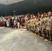 A military ceremony is taking place inside an auditorium or theater, with a formation of U.S. Army soldiers in OCP (Operational Camouflage Pattern) uniforms standing at attention along the right side. Civilian attendees, likely family members and guests, are standing on the left side of the venue. The setting suggests a formal military event such as a promotion ceremony, graduation, or unit activation/deployment ceremony.