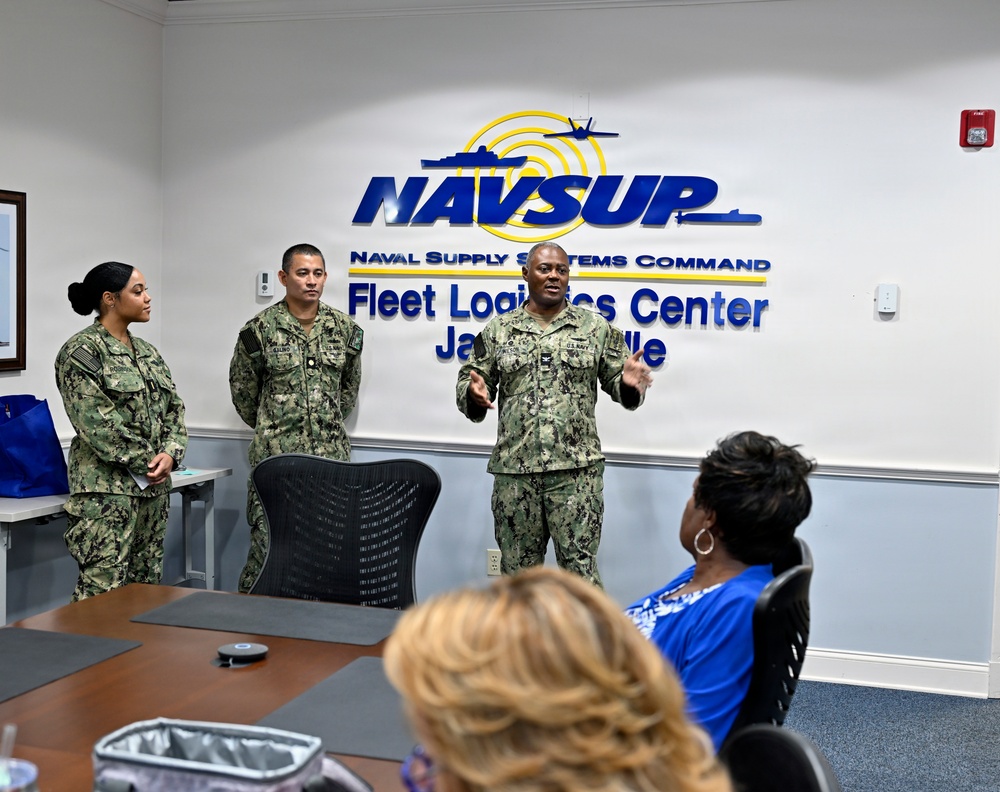 Three U.S. Navy personnel in NWU Type III (woodland digital camouflage) uniforms are presenting or briefing a small audience in a conference room at NAVSUP Fleet Logistics Center Jacksonville. One senior sailor appears to be speaking with open gestures while two others stand at attention beside him. Civilian attendees are seated at a conference table facing the presenters.