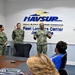Three U.S. Navy personnel in NWU Type III (woodland digital camouflage) uniforms are presenting or briefing a small audience in a conference room at NAVSUP Fleet Logistics Center Jacksonville. One senior sailor appears to be speaking with open gestures while two others stand at attention beside him. Civilian attendees are seated at a conference table facing the presenters.