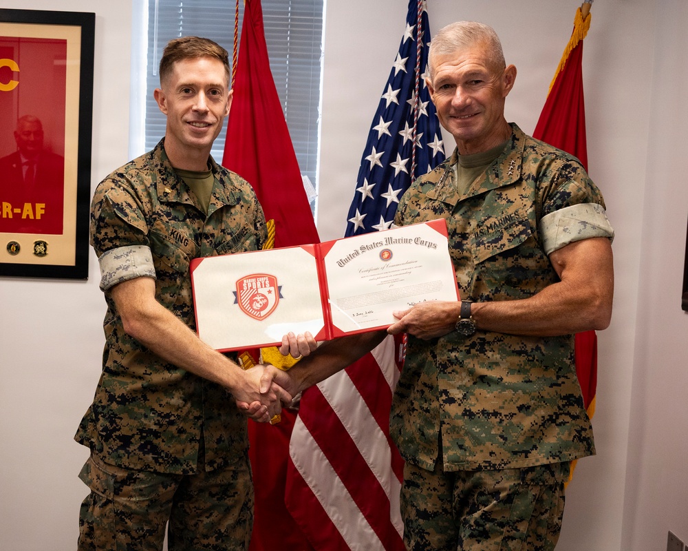 Two United States Marine Corps personnel in MARPAT camouflage uniforms are posing together while holding an open certificate folder displaying a 'United States Marine Corps Certificate of Commendation' with a Marine Corps Sports logo on the left panel. They are shaking hands in what appears to be an official award or recognition ceremony. The setting is an indoor office environment decorated with military flags and insignia.