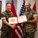 Two United States Marine Corps personnel in MARPAT camouflage uniforms are posing together while holding an open certificate folder displaying a 'United States Marine Corps Certificate of Commendation' with a Marine Corps Sports logo on the left panel. They are shaking hands in what appears to be an official award or recognition ceremony. The setting is an indoor office environment decorated with military flags and insignia.
