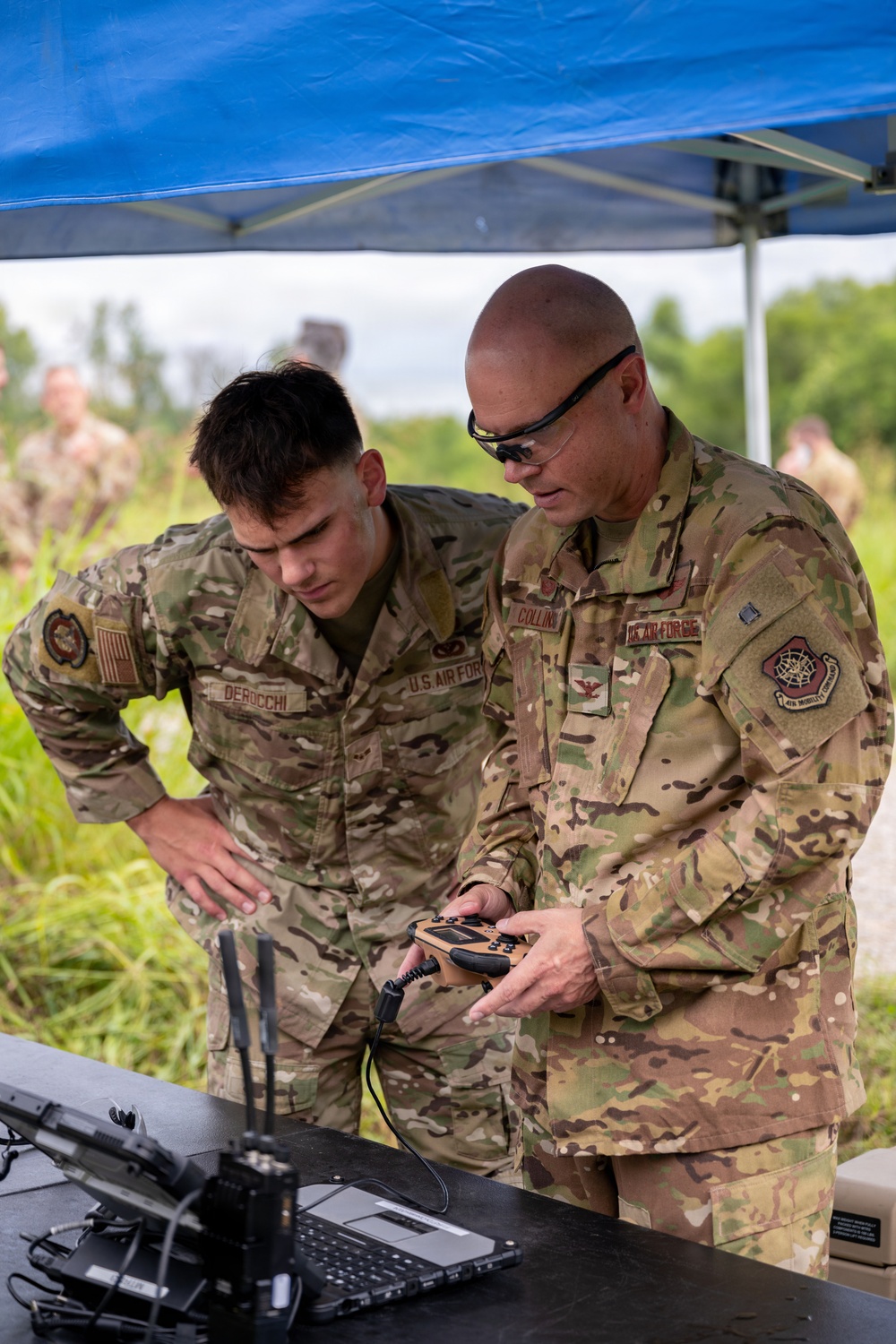 Two U.S. Air Force personnel in OCP (Operational Camouflage Pattern) uniforms are examining equipment at an outdoor field station under a blue canopy tent. The individual on the right, identified by name tape as 'COLLINS' and wearing a colonel's eagle insignia, holds a tan handheld device while both personnel review a laptop and associated equipment on a table. Additional military personnel are visible in the background in a grassy outdoor environment.