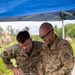 Two U.S. Air Force personnel in OCP (Operational Camouflage Pattern) uniforms are examining equipment at an outdoor field station under a blue canopy tent. The individual on the right, identified by name tape as 'COLLINS' and wearing a colonel's eagle insignia, holds a tan handheld device while both personnel review a laptop and associated equipment on a table. Additional military personnel are visible in the background in a grassy outdoor environment.