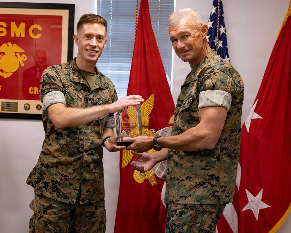 Two U.S. Marine Corps personnel in MARPAT camouflage uniforms are posing together while jointly holding a crystal/glass award trophy. The ceremony appears to take place in an official military office setting with USMC insignia and flags visible in the background. The individual on the right appears to be a general officer based on the star insignia visible on the flag beside him.