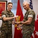 Two U.S. Marine Corps personnel in MARPAT camouflage uniforms are posing together while jointly holding a crystal/glass award trophy. The ceremony appears to take place in an official military office setting with USMC insignia and flags visible in the background. The individual on the right appears to be a general officer based on the star insignia visible on the flag beside him.