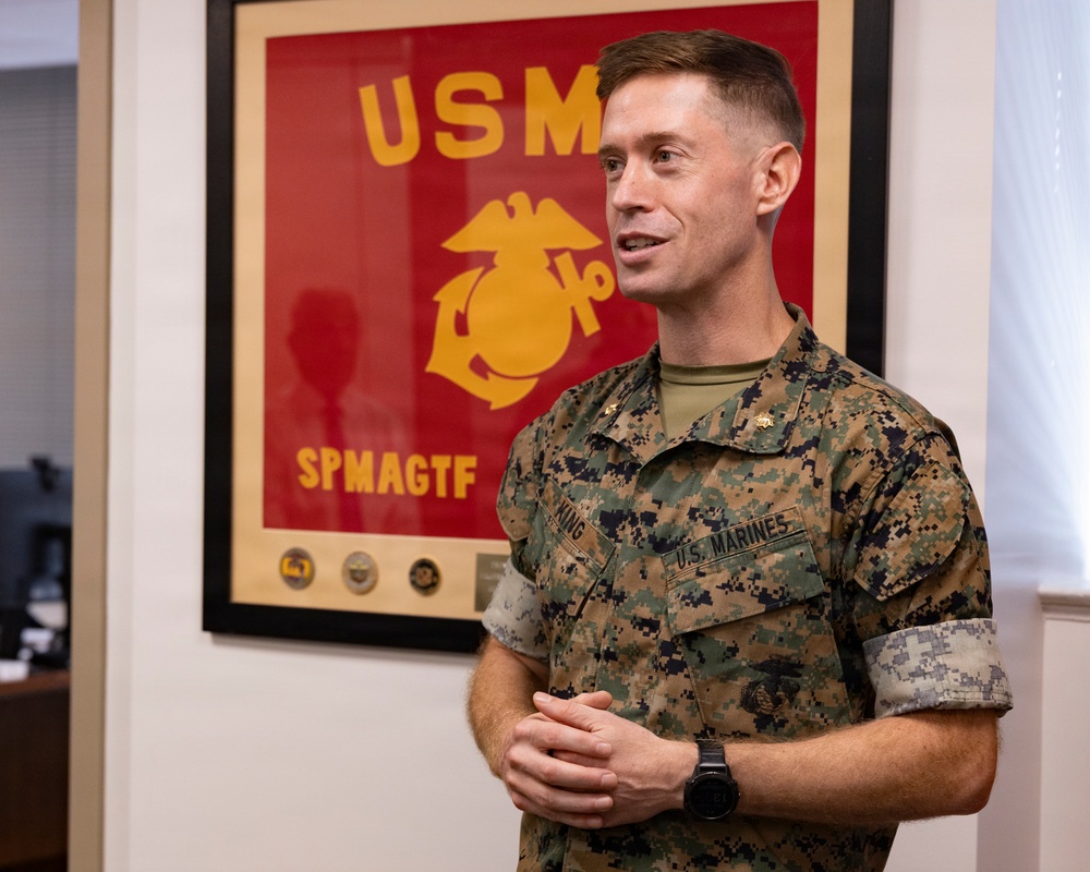 A U.S. Marine in MARPAT digital camouflage uniform stands in an indoor office setting, appearing to speak or brief someone. Behind him is a framed USMC unit flag/poster with the Eagle, Globe, and Anchor emblem and the text 'SPMAGTF' (Special Purpose Marine Air-Ground Task Force). His name tape reads 'KING' and he wears rank insignia consistent with a field-grade officer.