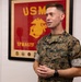 A U.S. Marine in MARPAT digital camouflage uniform stands in an indoor office setting, appearing to speak or brief someone. Behind him is a framed USMC unit flag/poster with the Eagle, Globe, and Anchor emblem and the text 'SPMAGTF' (Special Purpose Marine Air-Ground Task Force). His name tape reads 'KING' and he wears rank insignia consistent with a field-grade officer.