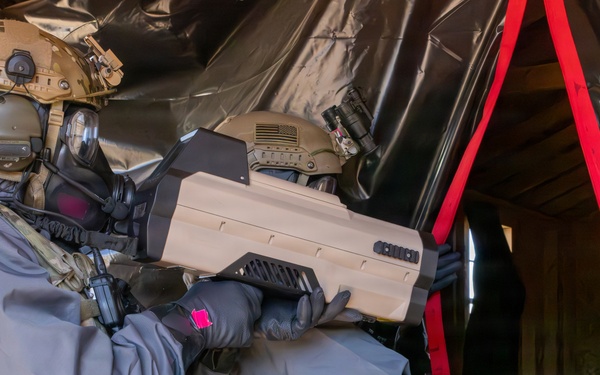 Two military or law enforcement personnel in full tactical gear, including gas masks and multicam uniforms, are conducting what appears to be a training exercise or operation inside a structure. One operator is holding a large tan-colored electronic device, possibly a counter-drone or electronic warfare system. The environment includes black plastic sheeting and red strapping, suggesting a controlled or improvised entry point.