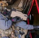 Two military or law enforcement personnel in full tactical gear, including gas masks and multicam uniforms, are conducting what appears to be a training exercise or operation inside a structure. One operator is holding a large tan-colored electronic device, possibly a counter-drone or electronic warfare system. The environment includes black plastic sheeting and red strapping, suggesting a controlled or improvised entry point.