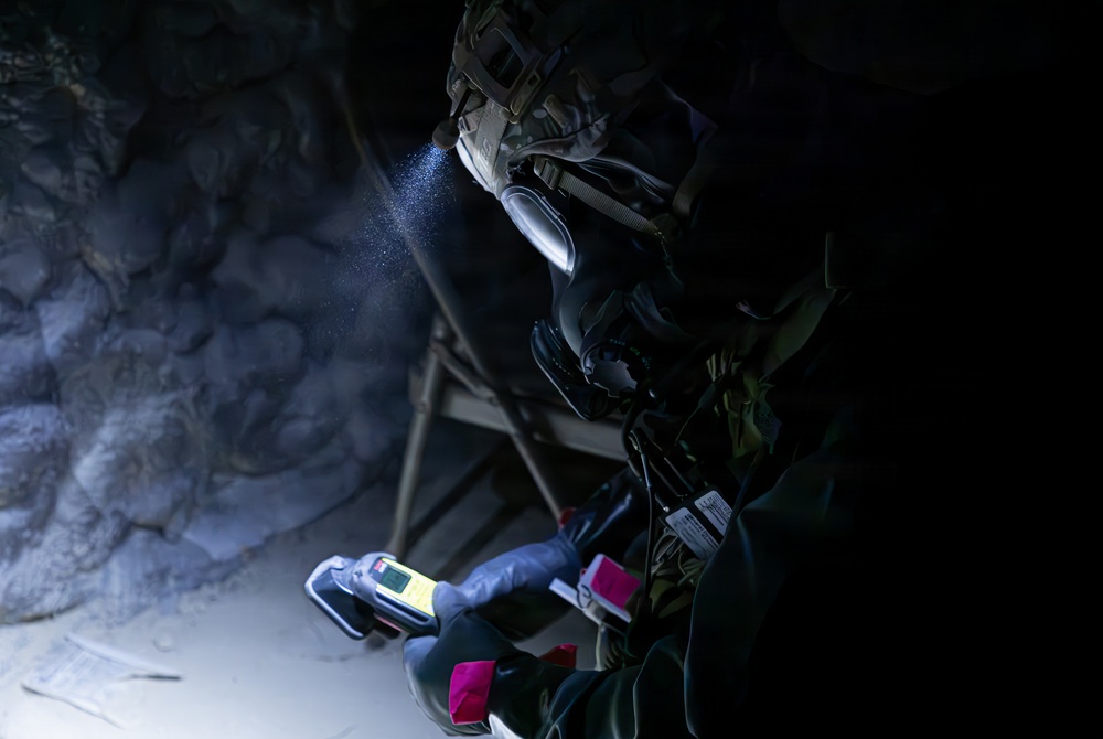 A person in military or tactical gear, wearing a full-face respirator mask and helmet with a mounted light, is operating inside a dark cave or tunnel environment. They are holding a handheld electronic detection device and what appears to be a chemical test kit or sampling material. The scene suggests a CBRN (Chemical, Biological, Radiological, Nuclear) reconnaissance or hazardous material assessment operation.